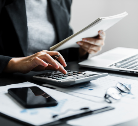 Eine Frau hält Papier in der Hand und sitzt an einem Schreibtisch mit Laptop, Taschenrechner, Smartphone und Dokumenten.