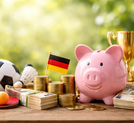 Rosa Sparschwein vor gestapelten Euro-Münzen und Geldbündeln, daneben ein Fußball, ein Tischtennisschläger, ein Federball und ein Pokal vor unscharfem grünem Hintergrund. Das Bild symbolisiert Fördermittel und finanzielle Unterstützung für Vereine.