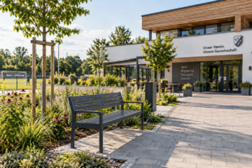 Ein gepflasterter Vorplatz eines modernen Vereinsheims mit einer dunklen Parkbank, jungen Bäumen, Staudenbeeten und einem Sportplatz im Hintergrund. Am Gebäude stehen Aufschriften wie „Unser Verein Unsere Gemeinschaft".
